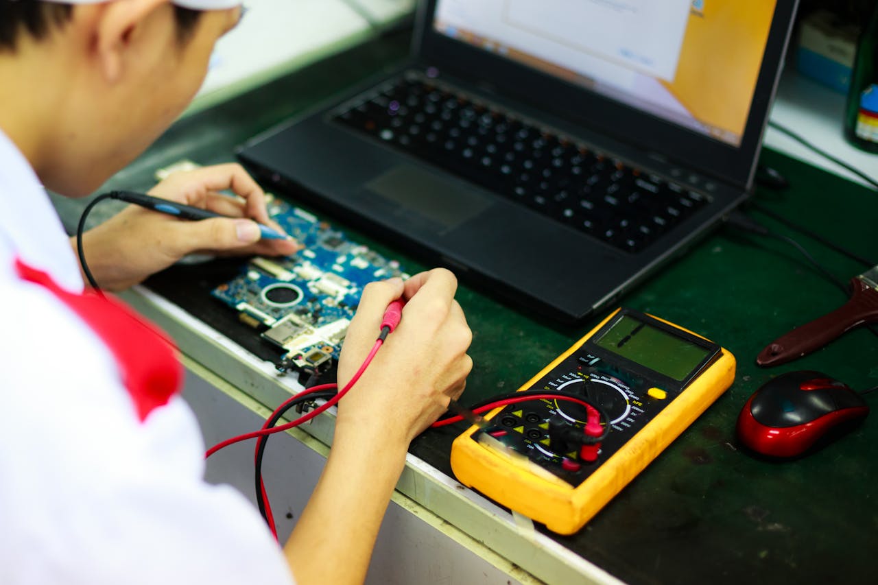 our-story Technician working on a laptop with multimeter in a professional workspace.